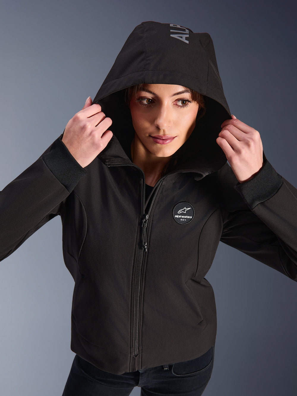 Woman wearing the Alpinestars Stella Radium Tech Hoodie in Black, urban motorcycle riding hoodie with hood up, featuring circular Alpinestars logo on chest, ribbed cuffs, and windproof 3-layer construction