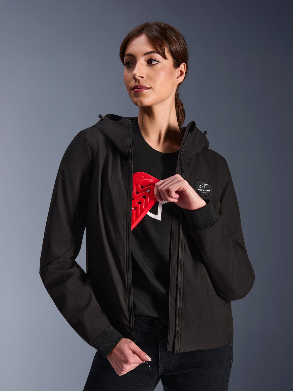 Alpinestars Woman Stella Radium Tech Hoodie, Black urban riding hoodie, female model demonstrating red Nucleon Flex Plus armor inside, lightweight 3-layer windproof textile shell with ribbed cuffs and chest logo patch, protective motorcycle apparel