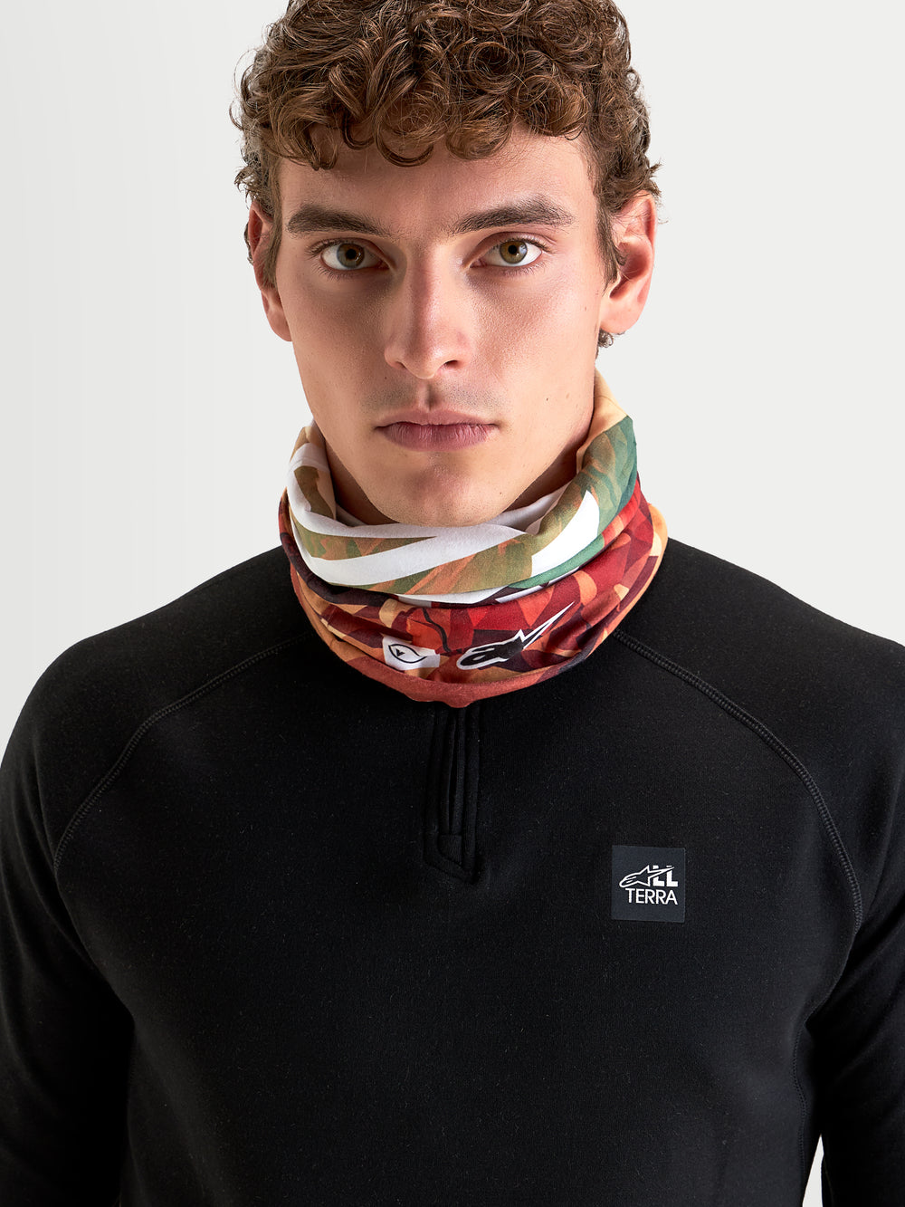 Alpinestars Buff Peaks Primaloft Neck Tube, earth brown and multicolored patterned neck gaiter, worn by a model in a black long-sleeve top, featuring low-bulk PrimaLoft insulation and seamless tubular construction for all-terrain motorcycle riding