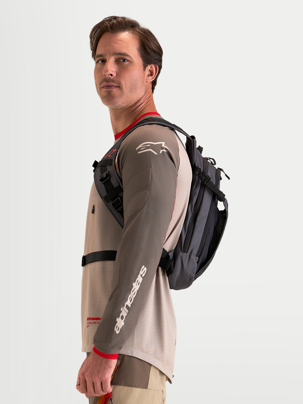 Alpinestars AT-9 Backpack, motocross backpack in walnut and deep gray, worn by a male model showing the side harness profile and lightweight textile construction