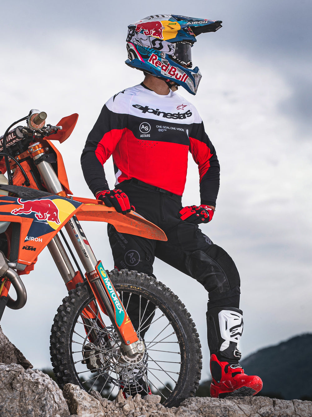 Motocross rider wearing Alpinestars Pro-Dura Jersey in Black, Bright Red, and White, standing next to a KTM dirt bike on rocky terrain, paired with black pants and red motocross boots, outdoor enduro setting.